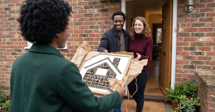 A friend in a green coat, seen from behind, hands a smiling couple a house-shaped photo collage at their open front door