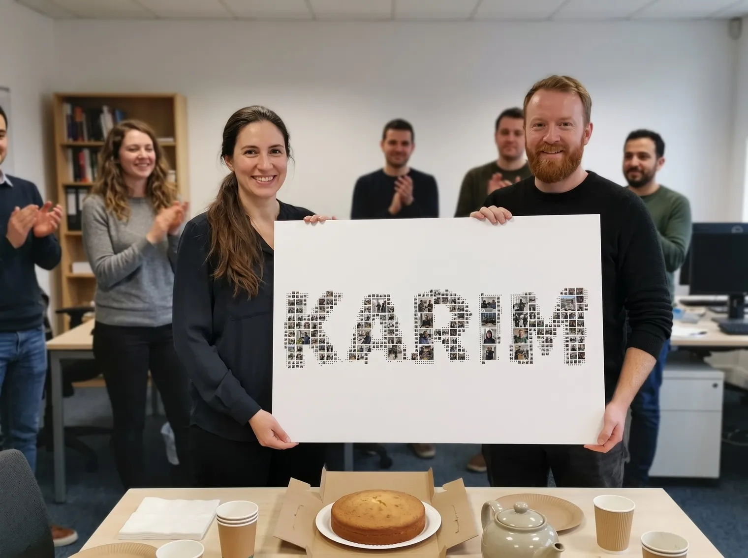 A woman with long dark hair in a ponytail and a man with a ginger beard hold up the KARIM name collage printed on a white foam board at a small office farewell, with a plain cake and tea in front